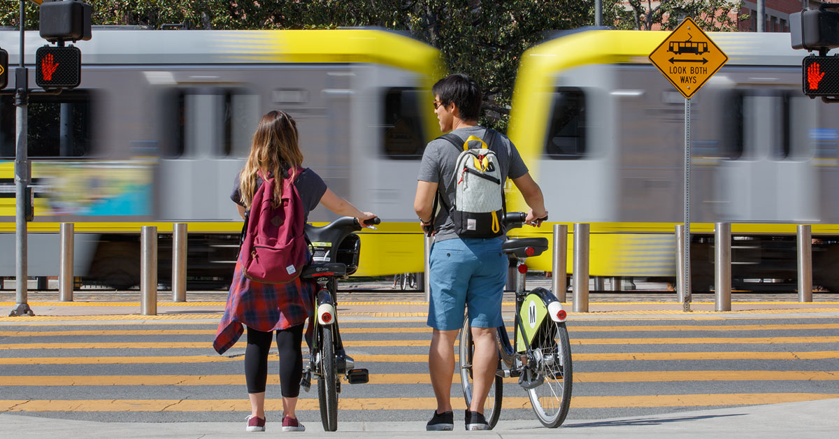 Two cyclists approaching a Metro Rail station with a train moving past in the distance.